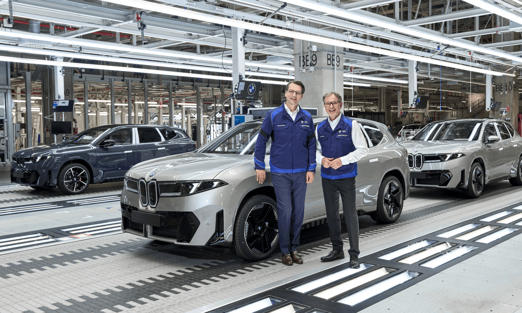 two men standing in front of a bmw in a factory